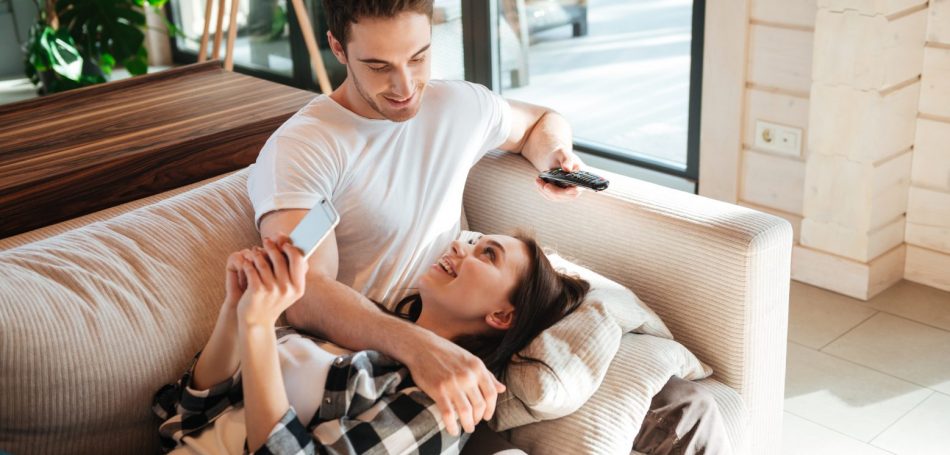 Talking smiling woman lying on couch with her man holding remote controoler in living room